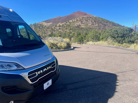 Silver Winnebago RV rental van on a winding road, facing a volcanic mountain with sparse trees under a blue sky. Great for travel!