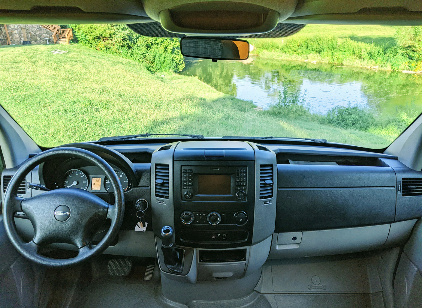 From inside an RV rental: driver's dash, steering wheel, and a vibrant view of green grass and a tranquil river.