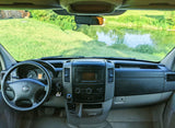 From inside an RV rental: driver's dash, steering wheel, and a vibrant view of green grass and a tranquil river.