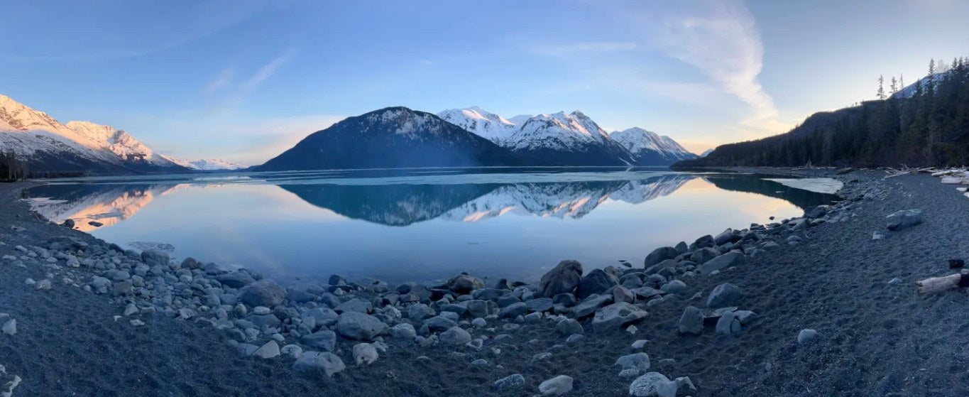 Panoramic view of a serene lake reflecting snowy mountains and a rocky shore, perfect for an RV rental trip.