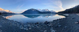 Panoramic view of a serene lake reflecting snowy mountains and a rocky shore, perfect for an RV rental trip.