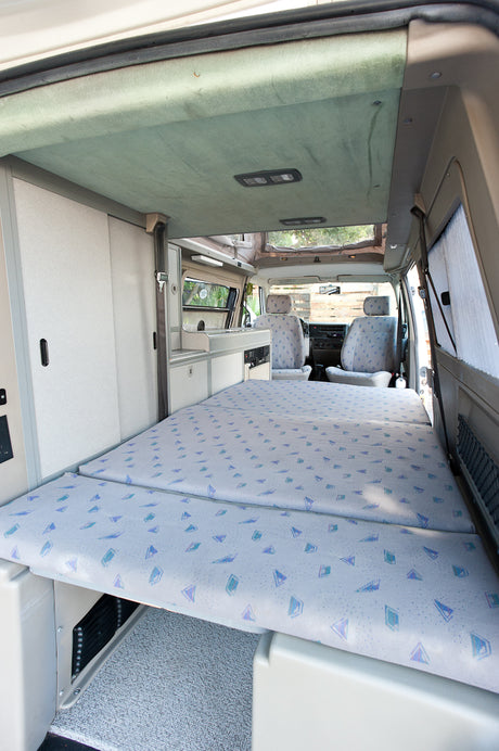 This RV rental camper van interior features a folded-out bed with patterned cushions, storage cabinets, and the front cockpit.
