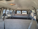 Interior of a passenger van, showing rows of grey fabric seats and hanging seatbelts. An RV rental and other vehicles are visible outside.