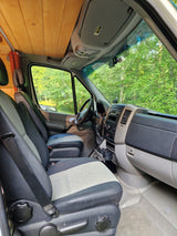 Front cabin of an RV rental: driver and passenger seats, dashboard, wooden ceiling, and green trees outside.