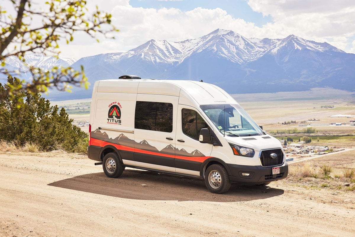 This white Ford Transit rv rental by Titus Adventure Co. is parked on a dirt road with snow-capped mountains and valley views.|This RV rental interior offers a raised bed