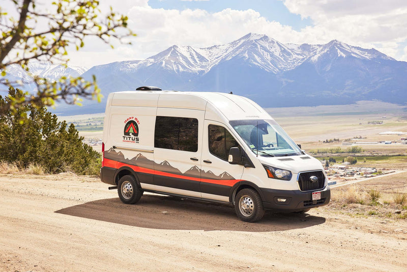 This white Ford Transit rv rental by Titus Adventure Co. is parked on a dirt road with snow-capped mountains and valley views.|This RV rental interior offers a raised bed — photo 1
