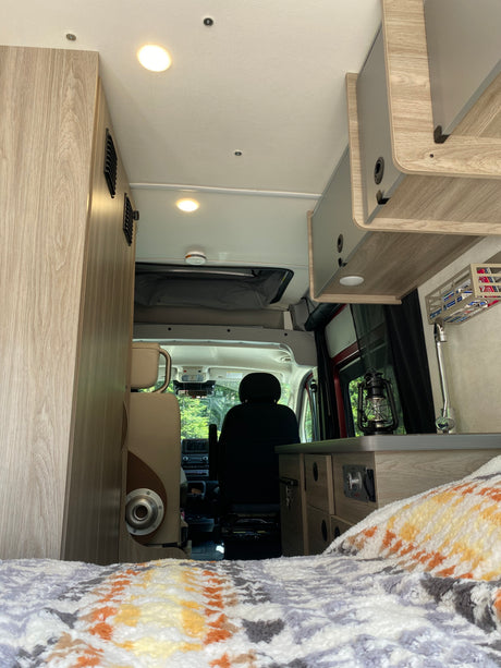 Cozy RV rental interior: light wood cabinetry, bed with colorful blanket, kitchen area, and driver's seat.