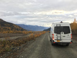 Adventurous rv rental: a white Sprinter van on a gravel road in Alaska's autumn mountains.