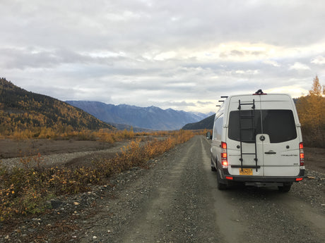 Adventurous rv rental: a white Sprinter van on a gravel road in Alaska's autumn mountains.
