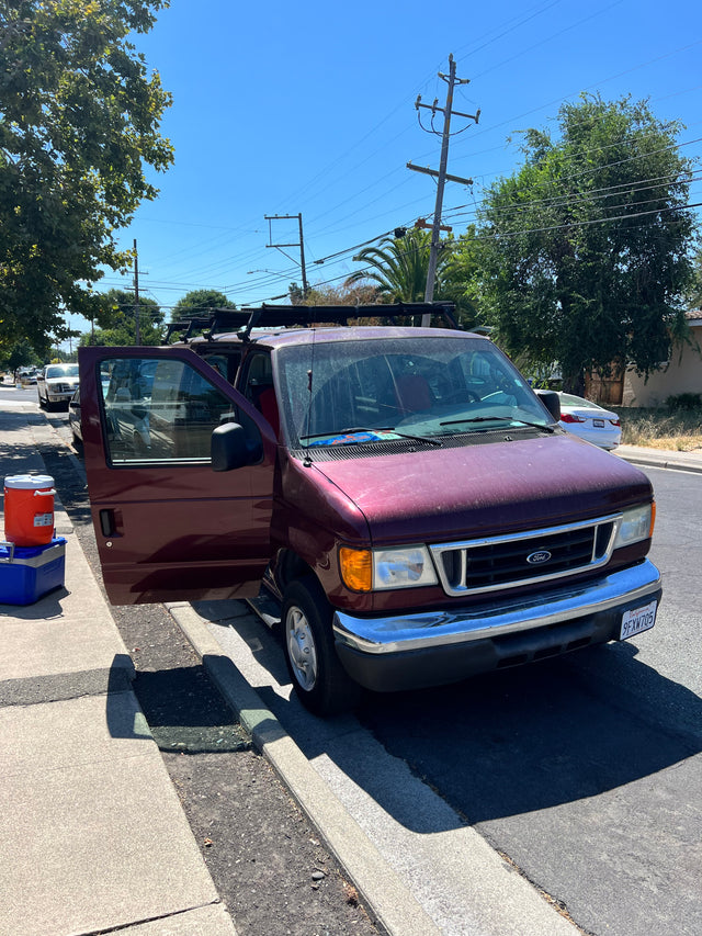 Maroon Ford van parked roadside with open door, coolers, and roof rack, possibly for RV rental adventures.