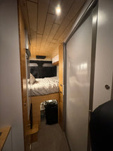 Inside an RV rental: a modern, narrow interior with a cozy bed, under-bed storage, wooden cabinetry, and a light sliding door.