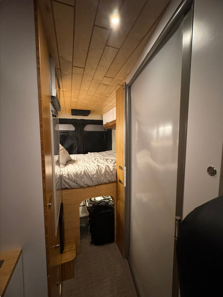Inside an RV rental: a modern, narrow interior with a cozy bed, under-bed storage, wooden cabinetry, and a light sliding door.