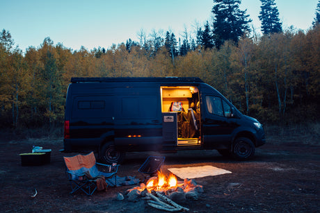 Cozy fall camping with an RV rental: a black camper van, glowing campfire, and comfy chairs set against vibrant autumn trees.