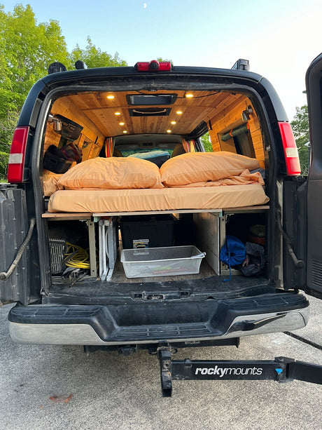 Back of a custom-built RV rental camper van, showing a cozy bed, wood ceiling, and under-bed storage.