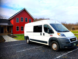 White Ram ProMaster camper van from our RV rental fleet, parked on a gravel lot beside a striking red building. Vermont views.