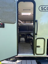 Open door of a light-colored rv rental, revealing a cozy interior with a bed, sofa, portable toilet, and skylight.