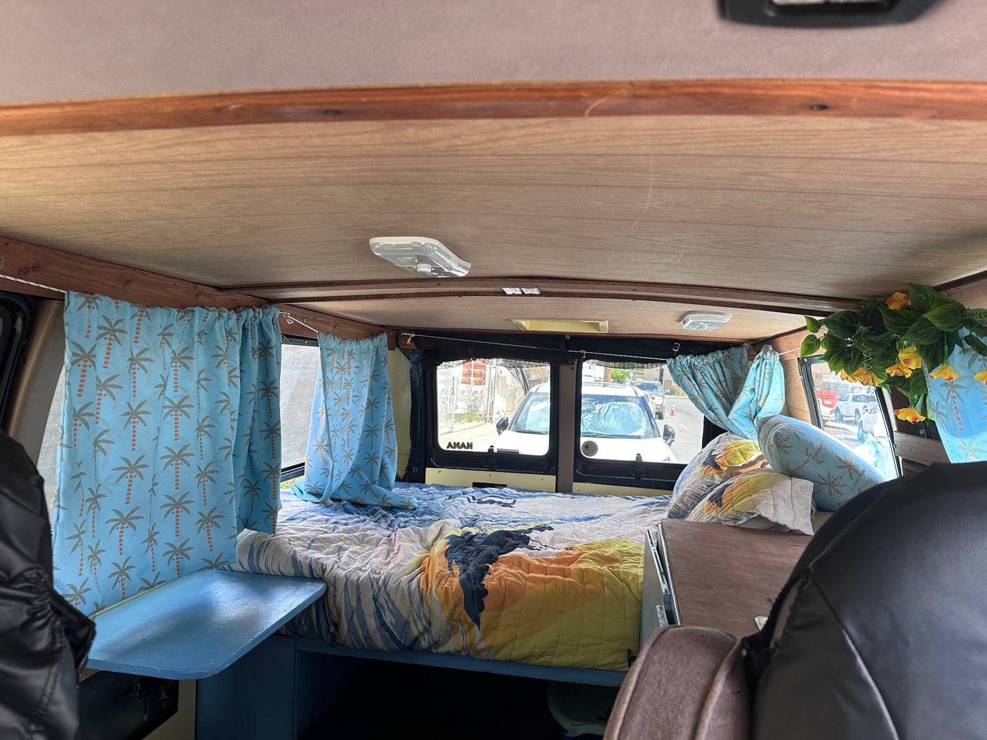 This RV rental interior features a bright bed, palm tree curtains, and wooden ceiling, ideal for comfortable campervan travel.