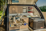 Compact RV rental's rear outdoor kitchen: stove, sink, and Coleman cooler setup for hassle-free camping cooking.