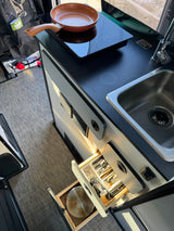 This rv rental features a compact kitchen with a sink, induction cooktop, and open drawers with utensils. A fire extinguisher is visible.
