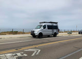 Silver Mercedes Sprinter camper van for rv rental, parked on a coastal road with ocean views under an overcast sky.