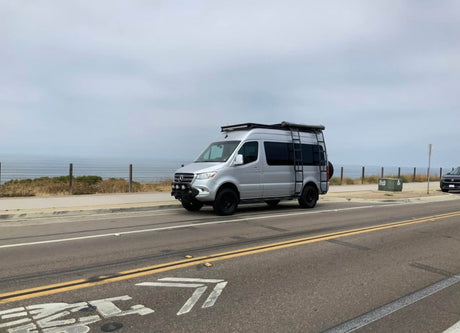 Silver Mercedes Sprinter camper van for rv rental, parked on a coastal road with ocean views under an overcast sky.