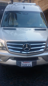 A silver Mercedes-Benz Sprinter van, often used for RV rental, front view with CA license plate 7GHX324. Palm trees reflect on it.