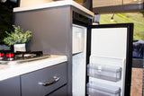 Inside an RV rental: a modern, compact kitchen with a gas stove, open refrigerator, and light grey cabinetry.
