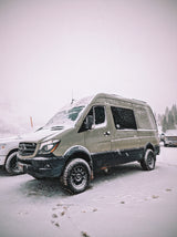 An olive green Mercedes Sprinter RV rental, fitted with large off-road tires, stands ready in a snowy winter landscape.