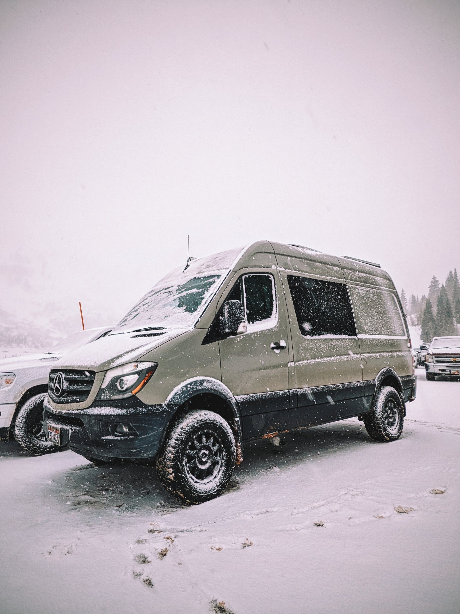 An olive green Mercedes Sprinter RV rental, fitted with large off-road tires, stands ready in a snowy winter landscape.