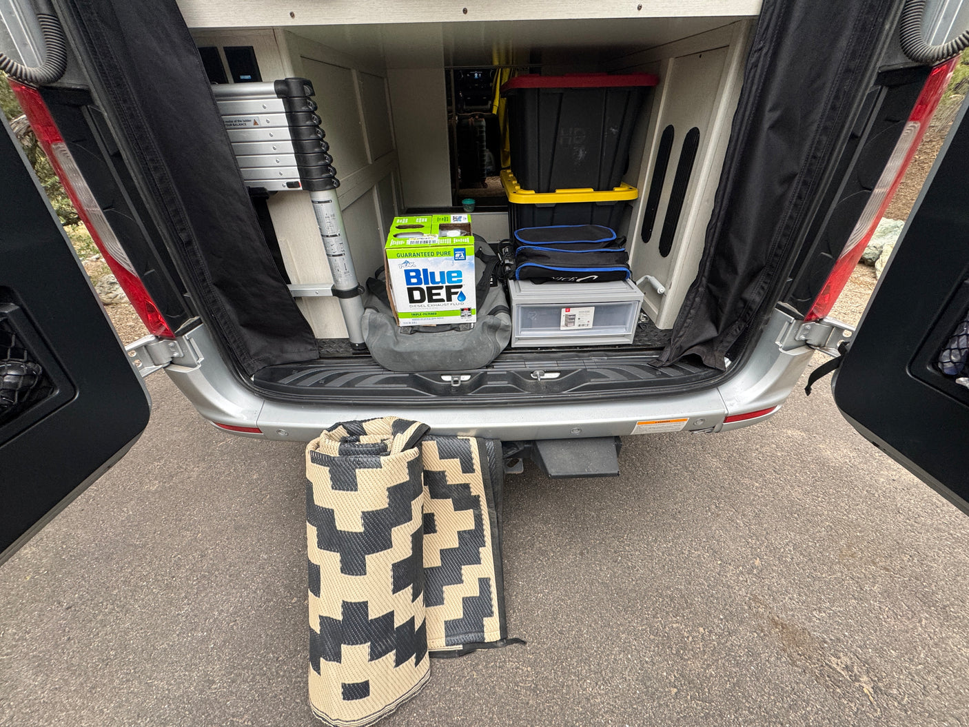 Open rear doors of an RV rental reveal organized camping gear: DEF, ladder, bins, & rolled mats.