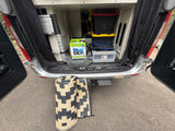 Open rear doors of an RV rental reveal organized camping gear: DEF, ladder, bins, & rolled mats.