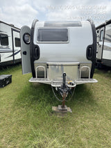 A sleek silver travel trailer, ready for RV rental, is parked on a grassy lot with other camping vehicles nearby.