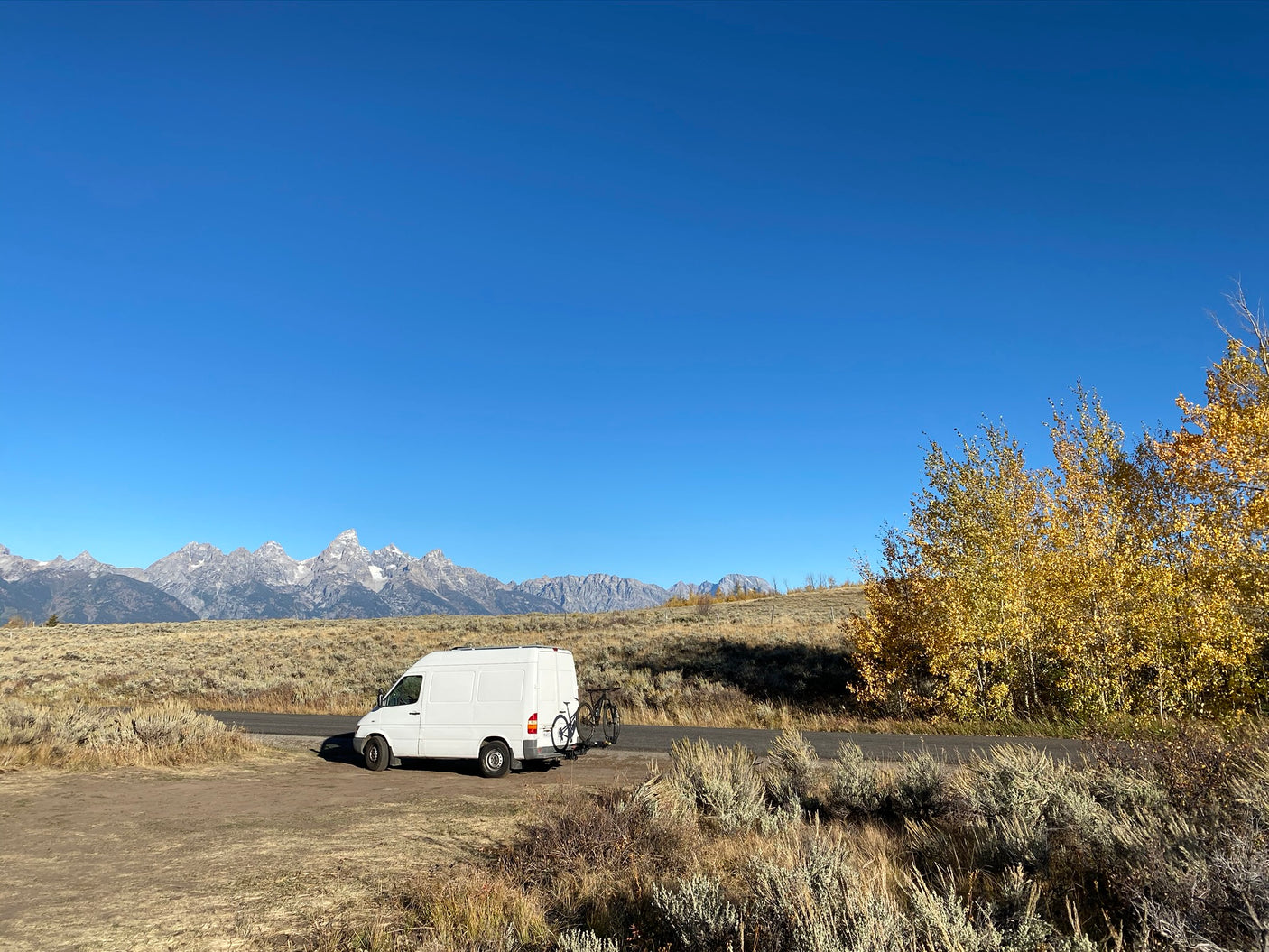 White RV rental van with bikes parked, overlooking majestic mountains and vibrant fall foliage under a clear blue sky.