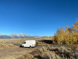 White RV rental van with bikes parked, overlooking majestic mountains and vibrant fall foliage under a clear blue sky.