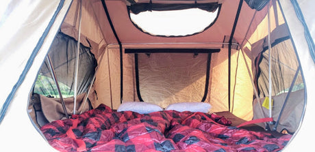 Cozy interior of an RV rental rooftop tent. Features a comfortable bed with red and black plaid bedding, metal poles, and mesh windows.