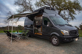 Dark gray Sprinter RV rental set up for outdoor dining. An extended awning covers a table with plates, wine, and chairs.