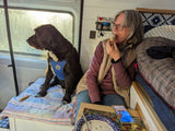 Inside an RV rental: a woman eats, a dog looks out a sunny window. Cozy space for travel with pets.
