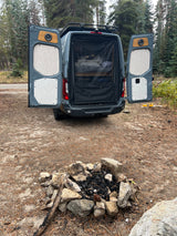 Ready for adventure! Rear view of an rv rental camper van with open doors, comfy bed, and a ready fire pit in the forest.