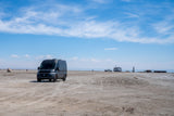 A dark grey camper van is parked on a vast, sandy area. In the distance, an RV rental is by the water under a blue sky.
