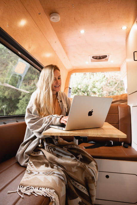 A woman works on her laptop inside a cozy RV rental, wrapped in a blanket, enjoying remote work amidst nature views.