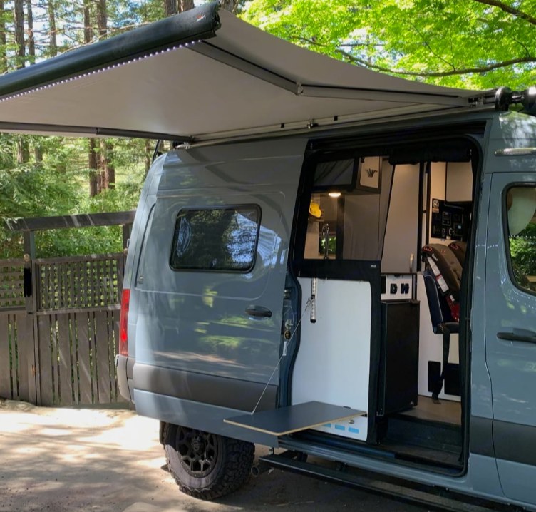 modern dashboard with a Mercedes steering wheel and comfy front seats.|Relax in this cozy RV rental bed. Enjoy the outdoor view & sunlight from your mobile bedroom retreat.|RV rental: Modern camper van interior with comfy bench seating — photo 4