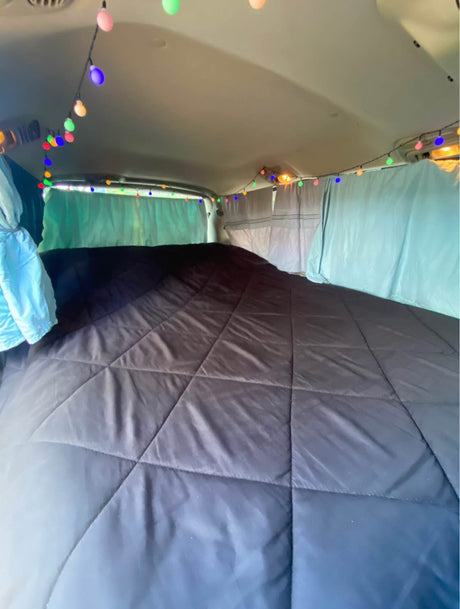 Cozy RV rental interior with a dark quilted bed, blue curtains, and colorful string lights for a comfortable road trip.