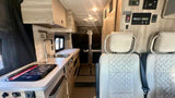 Modern RV rental interior: Light passenger seats, a compact kitchen with sink/stovetop, and wall-mounted control panel, looking to the rear.