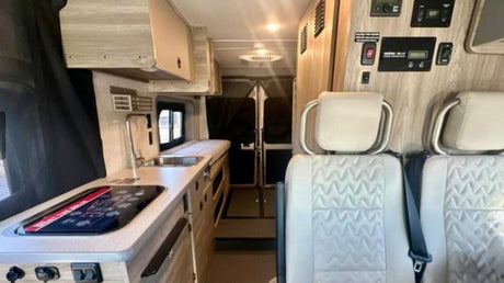 Modern RV rental interior: Light passenger seats, a compact kitchen with sink/stovetop, and wall-mounted control panel, looking to the rear.