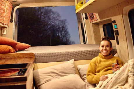 Smiling woman relaxes in a cozy RV rental camper van at night, enjoying the view from the open back.