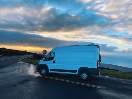 An rv rental camper van is parked on a wet, winding road at sunset, with a stunning ocean and dramatic sky view.