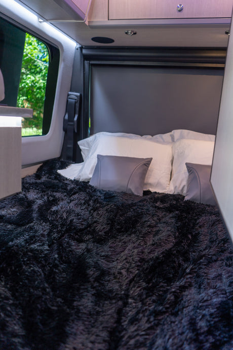 Explore in comfort! This RV rental's cozy sleeping area features a plush black blanket, pillows, and a scenic window view.