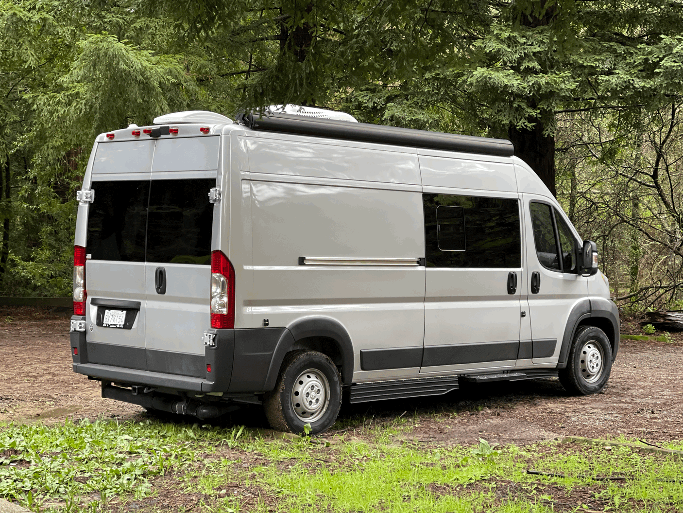 A silver camper van, ideal for RV rental, is parked outdoors. Features include a roof AC unit and awning, surrounded by green trees.