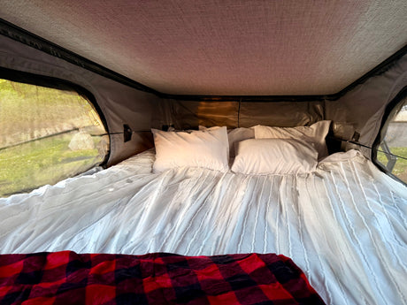 A comfy bed with white bedding & a red plaid blanket inside an RV rental tent, offering views of green outdoors.