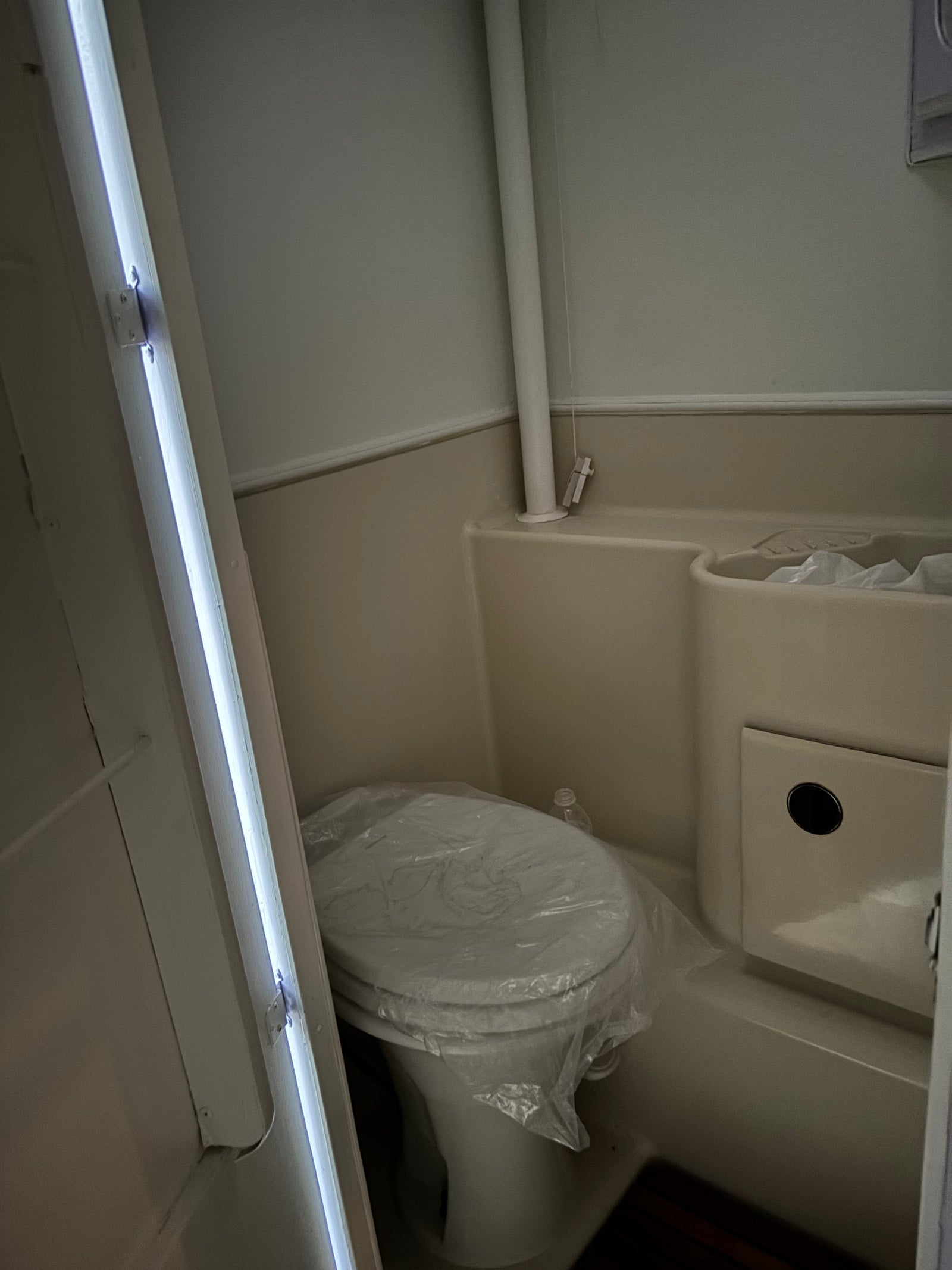 Compact RV rental bathroom with a new toilet (covered in plastic), integrated sink/shower, and bright vertical lighting.